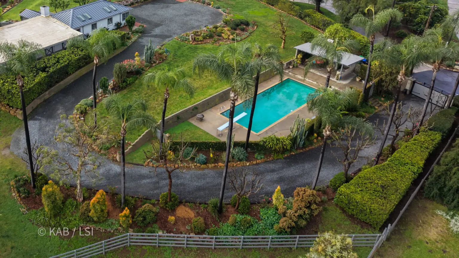 A private hillside pool terrace ringed by tall palms, with a guest house, looping gravel drives, and layered garden beds on a La Habra Heights property.