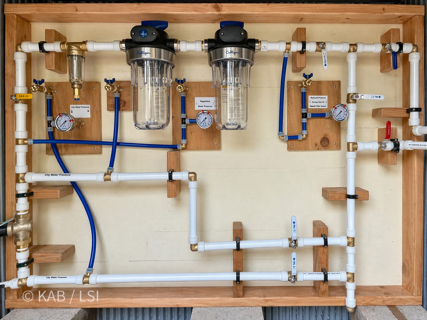 One-inch PEX whole-house water board built on the bench in Keith Bennett's Water House prototype: pressure regulator on left, SimPure spin-down sediment strainer, Culligan paper-cartridge filter, PurePlus activated carbon block, potassium softener branch, and a branch carrying full 120 PSI underground in 1 inch PVC to fire stations, RV pad, and home-office pad