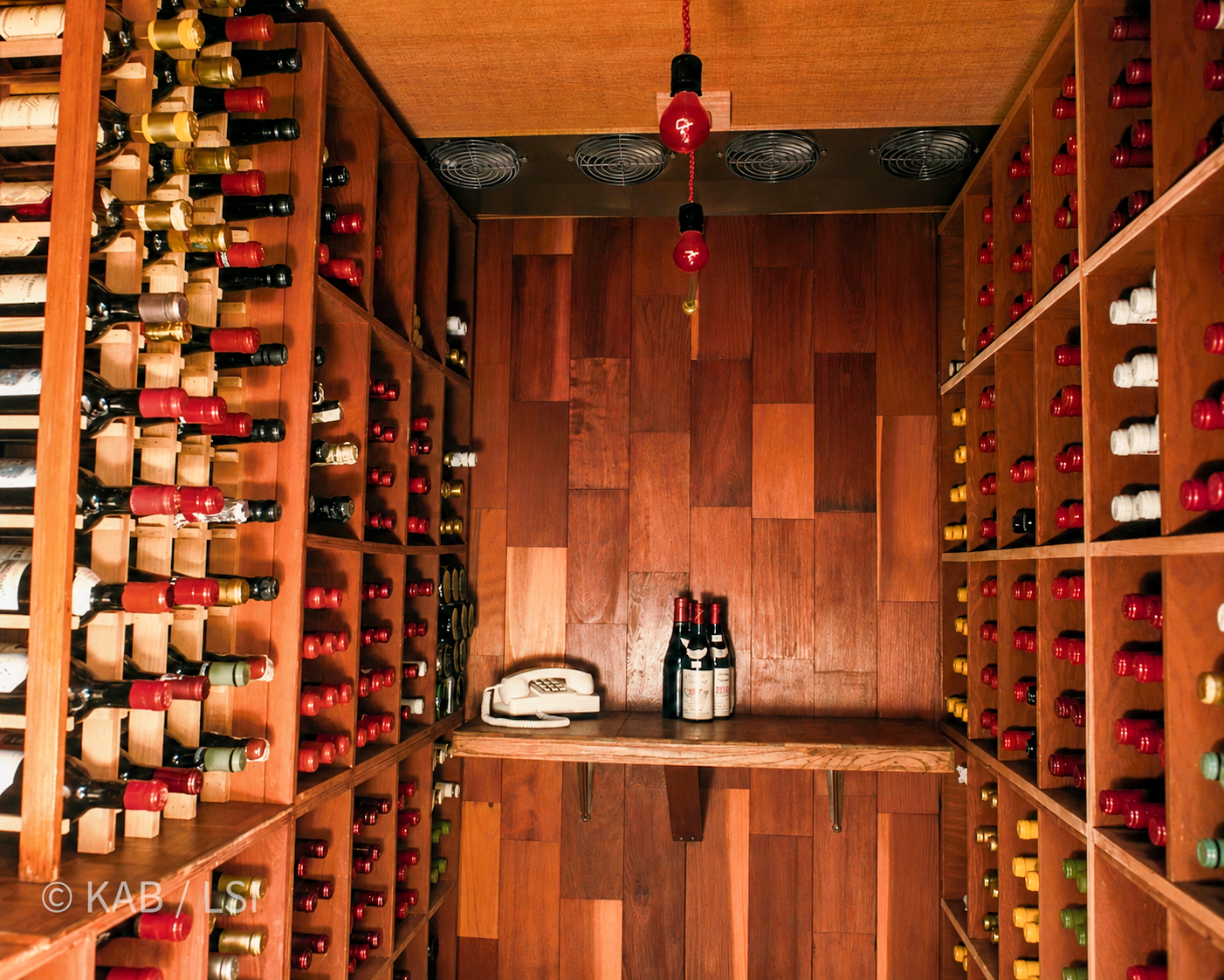 A finished wine cellar interior with full redwood racking, hundreds of bottles, a four-fan refrigeration array overhead, and a corded telephone on the uncorking shelf.