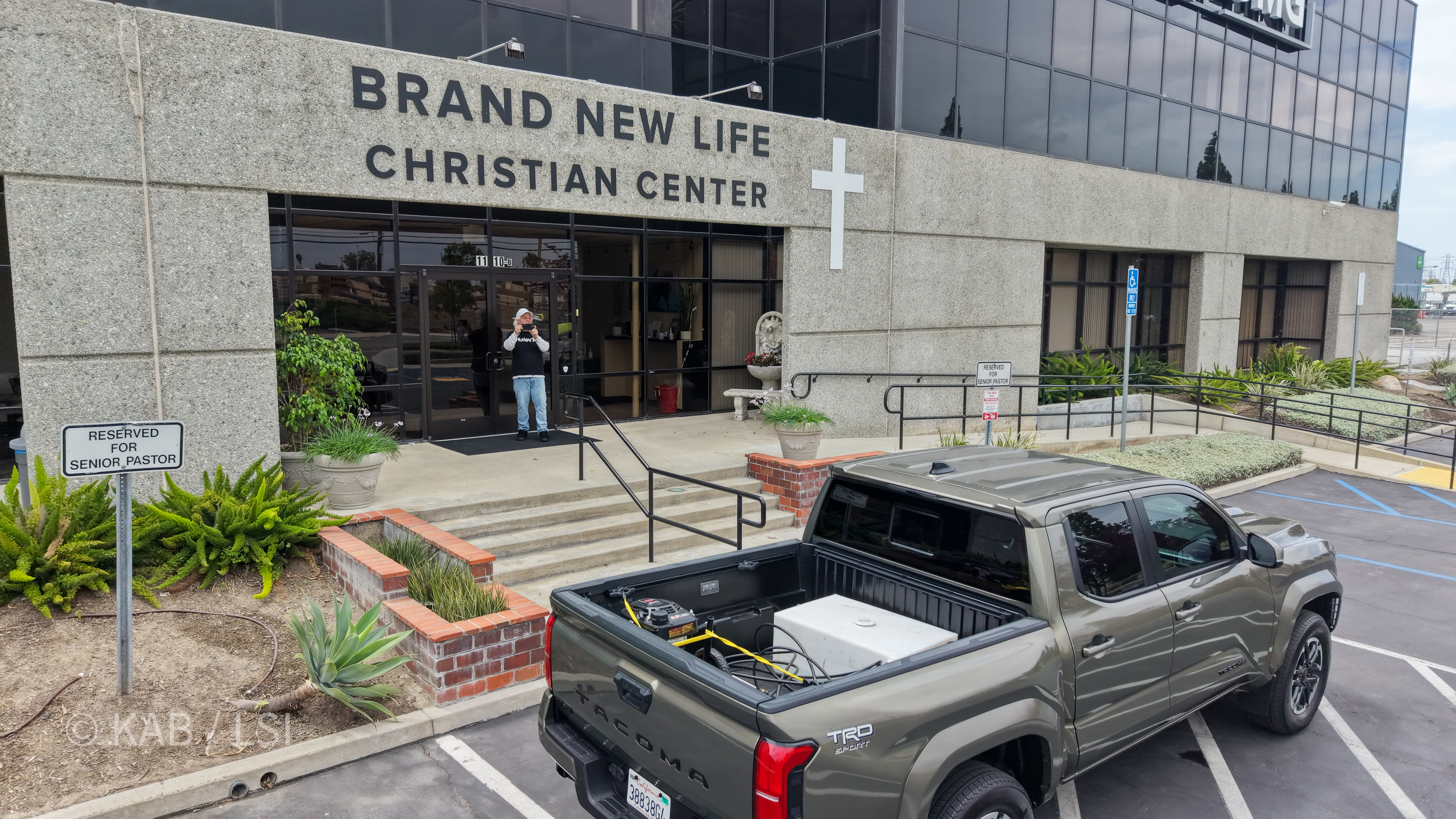 Front entrance of Brand New Life Christian Center at 1110 W. Taft Avenue, Orange, California, with the church's full name and cross on the building's stone facade. Longtime church volunteer Jeff Billig stands at the doorway in the foreground after locking up, photographing the drone overhead. A Reserved for Senior Pastor parking sign and ornamental aloe planter in the foreground.