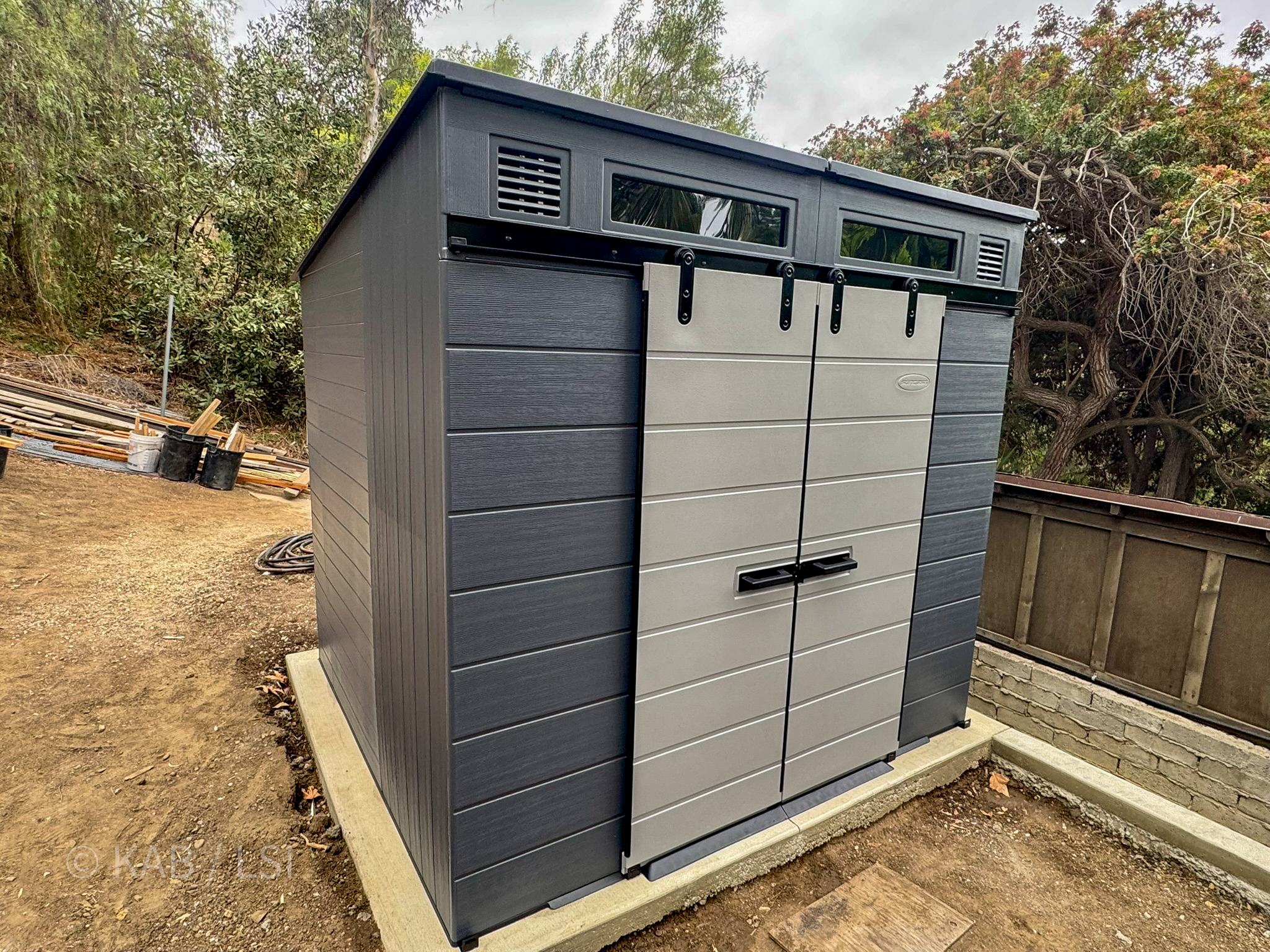 A pre-fab gray-and-slate plastic shed assembled on a fresh concrete slab — a tidy, modest outbuilding ready for use, surrounded by hillside earth and tools mid-job.