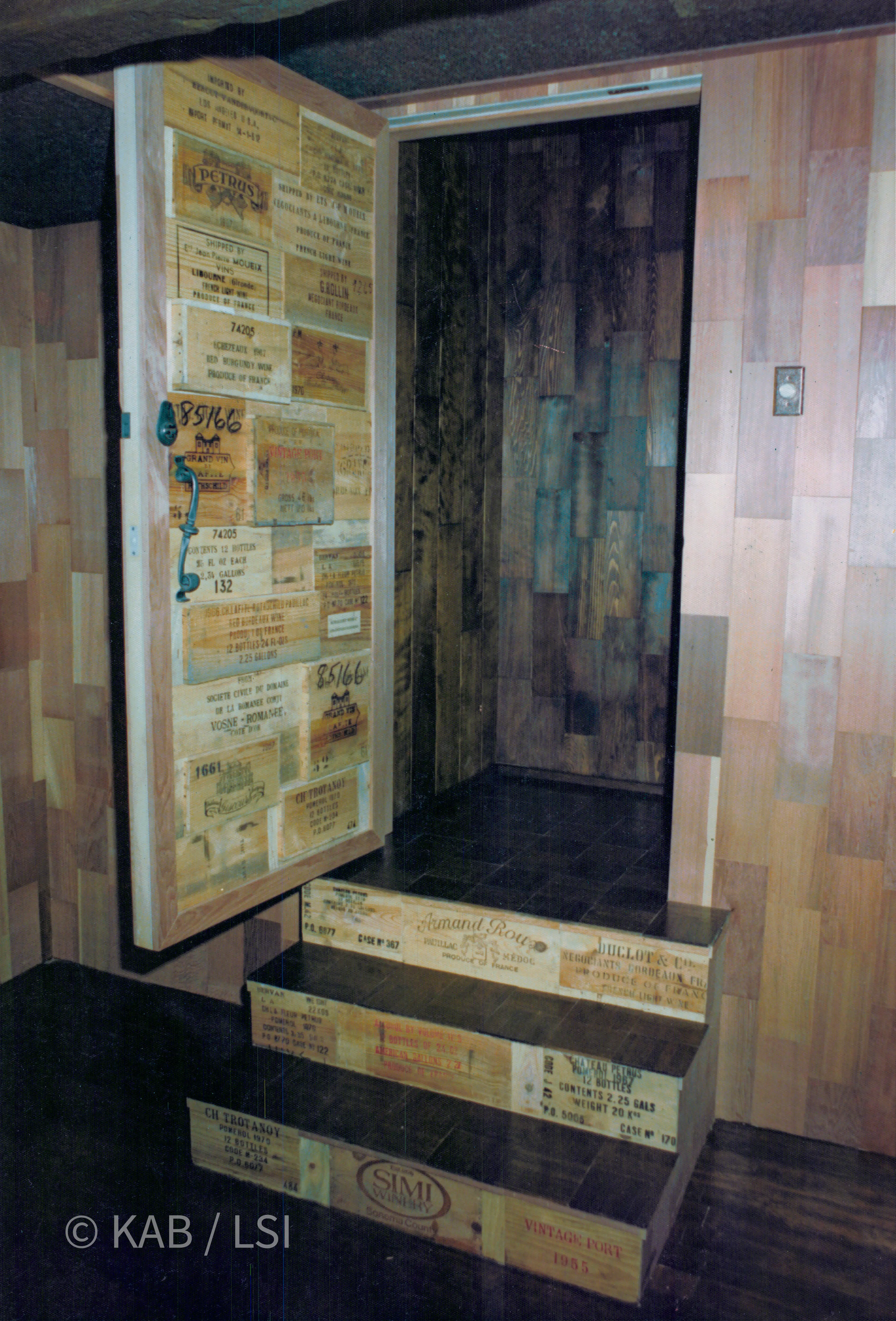A hidden cellar door faced entirely in stenciled wine-crate panels, standing open at the top of three crate-clad steps, with a redwood-lined cellar beyond.