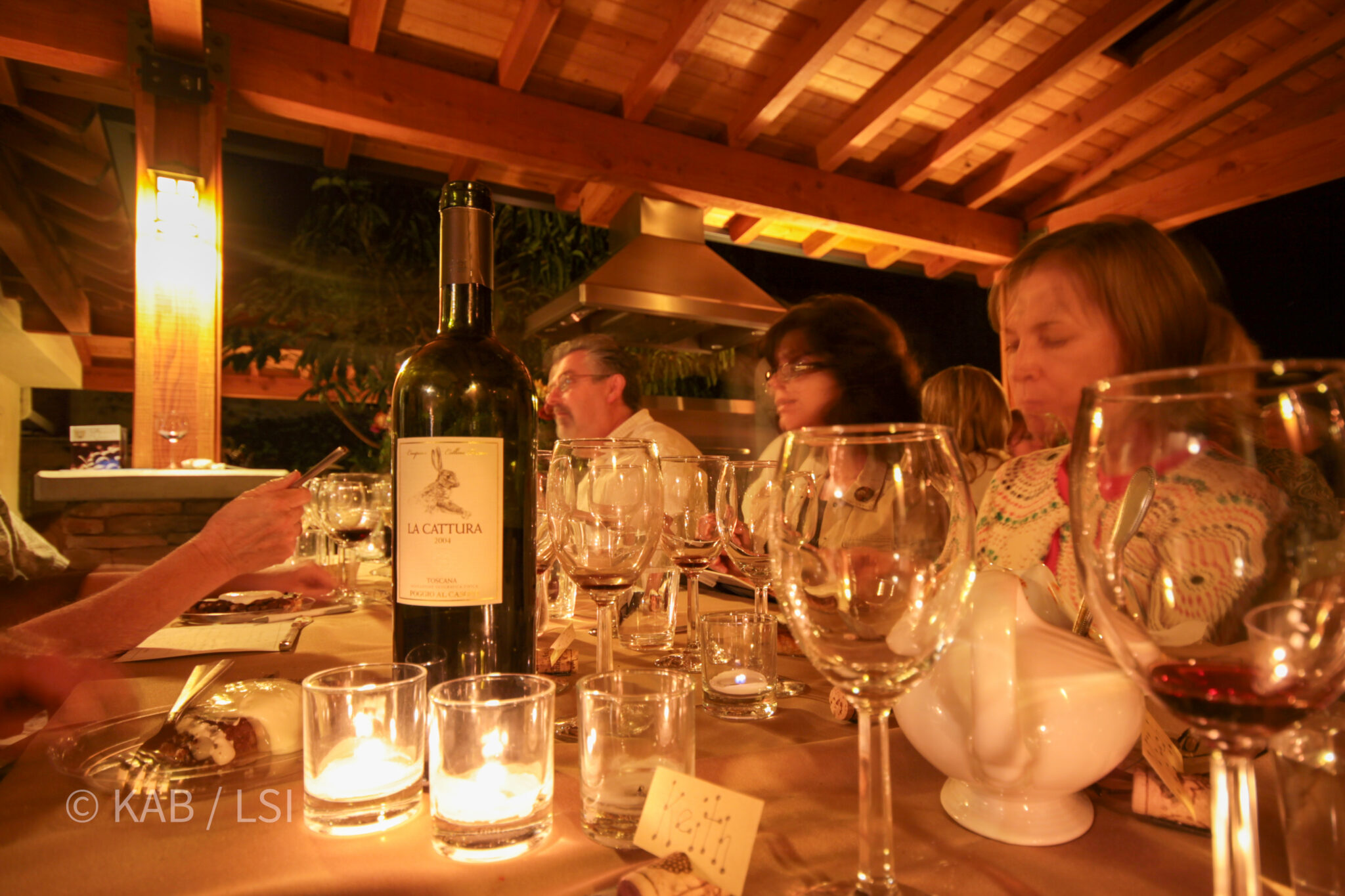 Guests gathered around a long candlelit table under a wood-beamed pergola at night, wine glasses and a standing bottle of red on the table.