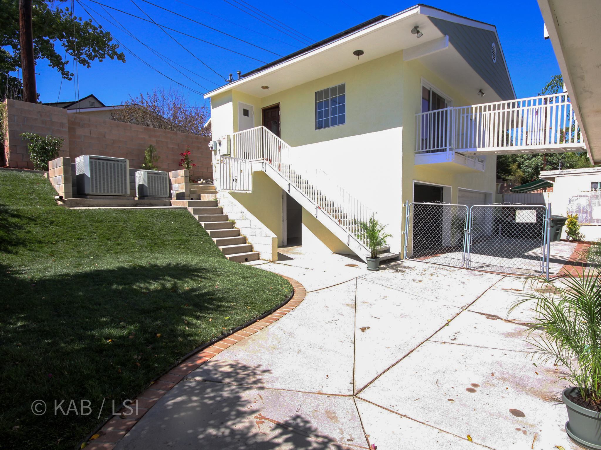 Side view of the completed La Mirada ADU and three-car garage built into the rear hillside cut. Yellow stucco main house at right with the new ADU box rising above the garage at center. Concrete-walled garage on the lower level, exterior staircase climbing to the ADU entry, central HVAC condensers staged on a curbed platform at left. Forty-year-old hillside-cut work, photographed after a full-property cleanup.