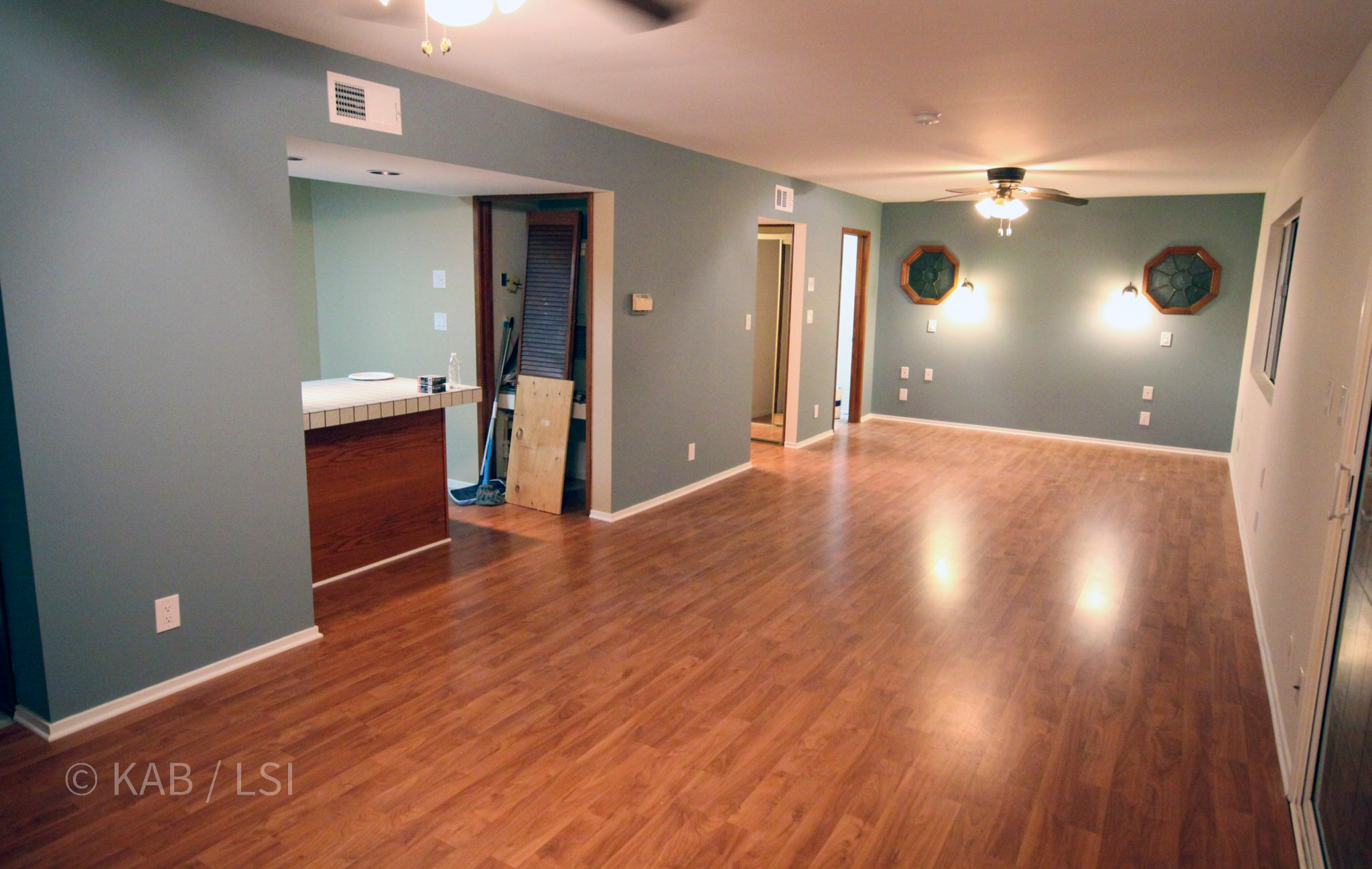 Interior of the completed 800-square-foot La Mirada ADU above the new three-car garage. Empty living and dining area photographed before move-in: sage-blue walls, hardwood floors, ceiling fan, two octagonal-frame wall sconces, kitchen pass-through at left, two bedroom doorways at center-right. The ADU includes a full kitchen, full bath, two walk-in closets, and a private patio bridged to the main house.