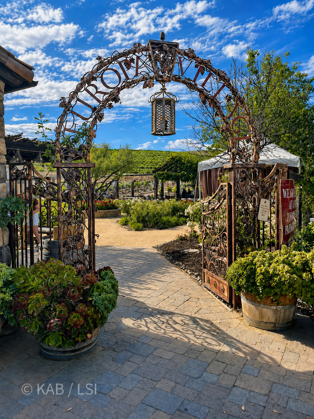 A wrought-iron entrance archway with grape-vine scrollwork and a hanging lantern, opening onto a winery garden and vineyard under a blue sky.