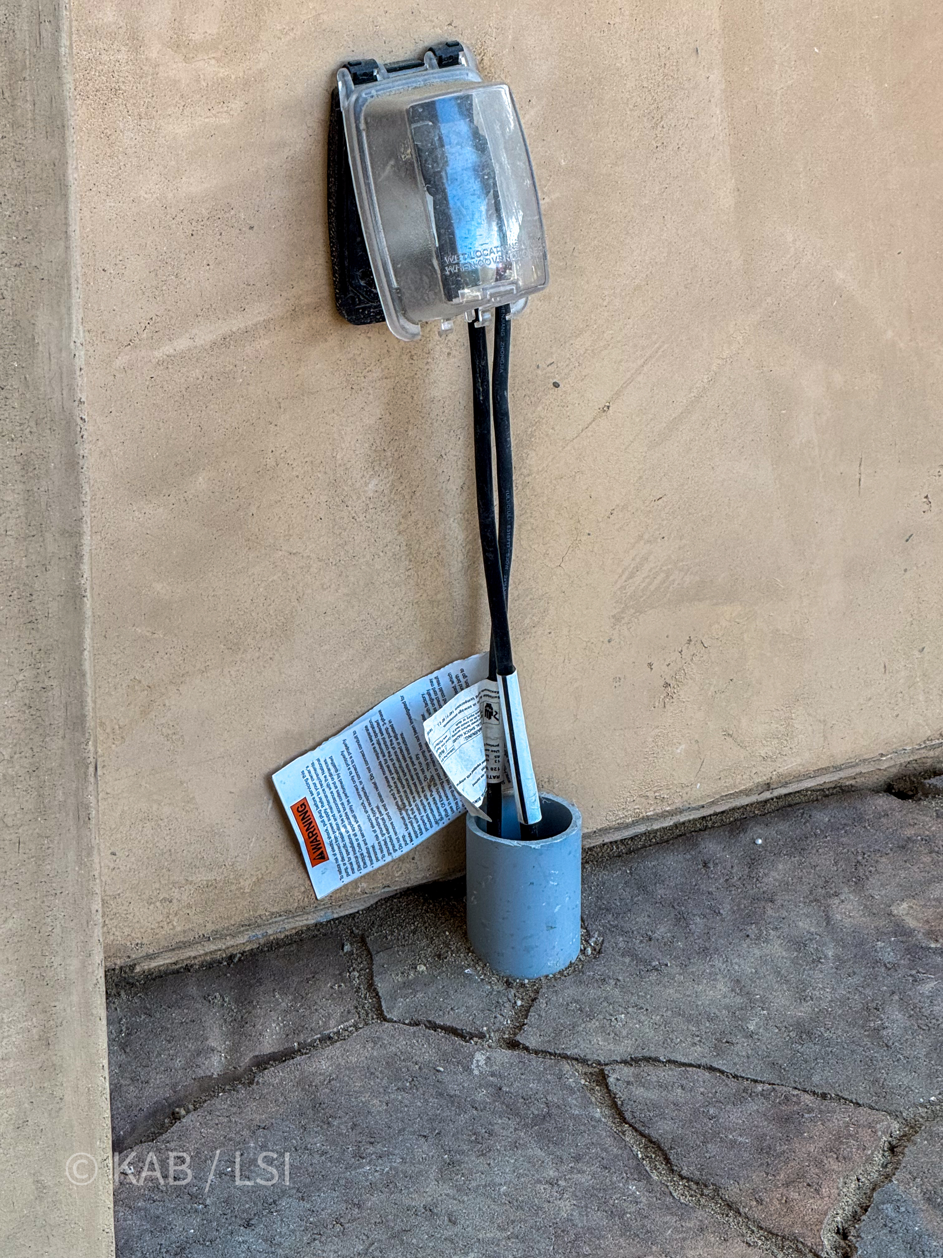 Photograph of an outdoor weather-resistant clamshell receptacle on a stucco wall, with two black industrial-strength extension cords running from the outlet down through a vertical gray PVC sleeve set into broken flagstone hardscape.